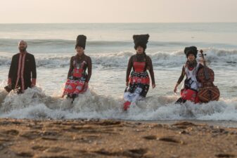 Photo of the four musicians in DakhaBrakha standing in the water, with the waves crashing up behind them.
