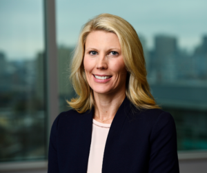 Professional portrait of a woman with blonde hair, styled in soft waves, wearing a navy blue blazer over a beige top. She is smiling, standing in front of a blurred background that appears to be an indoor staircase.