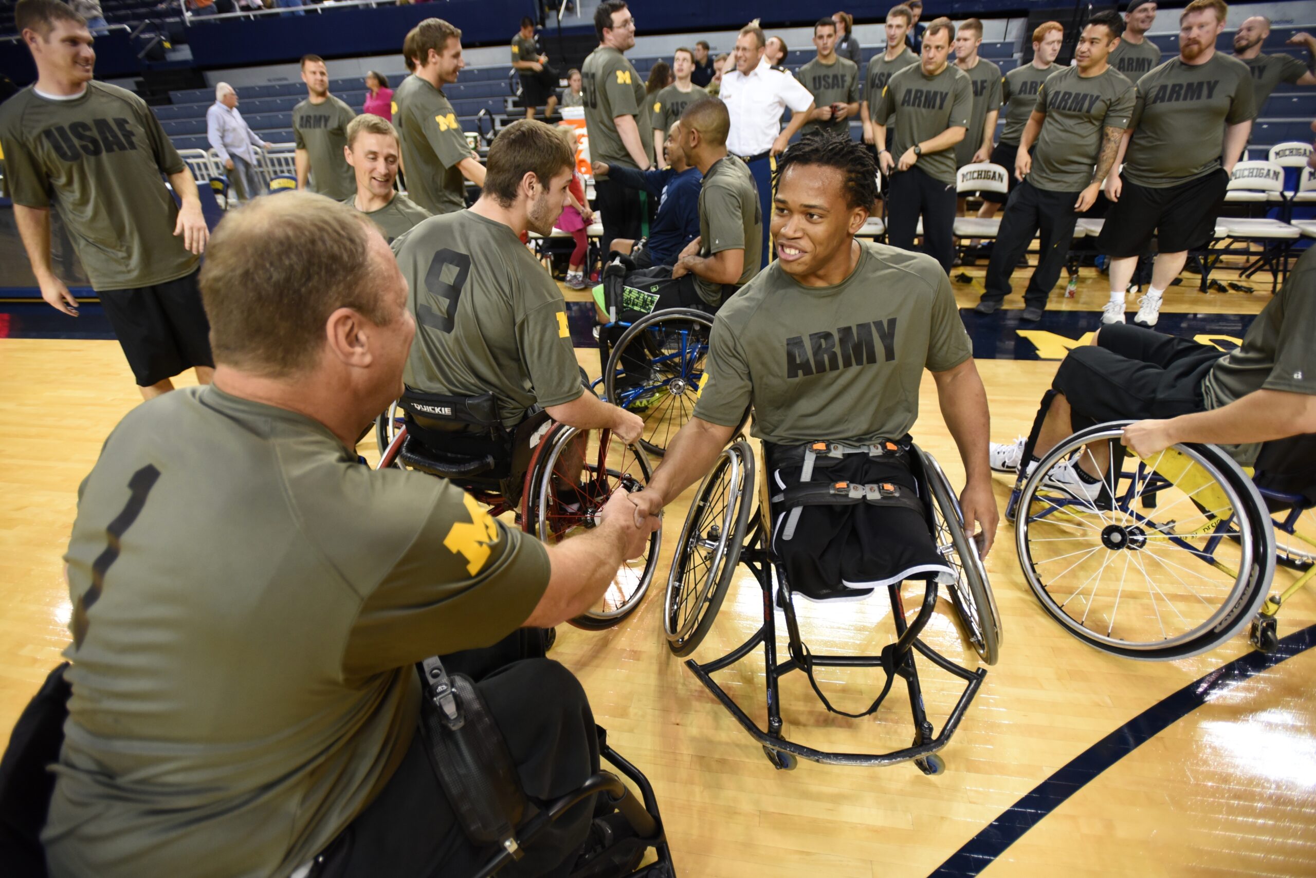 Players from Army and Air Force wheelchair basketball teams share a handshake and smiles on the court, demonstrating camaraderie and sportsmanship. The teams, wearing matching military-themed jerseys, are gathered in a circle with other players and supporters in the background, creating a powerful moment of unity and resilience