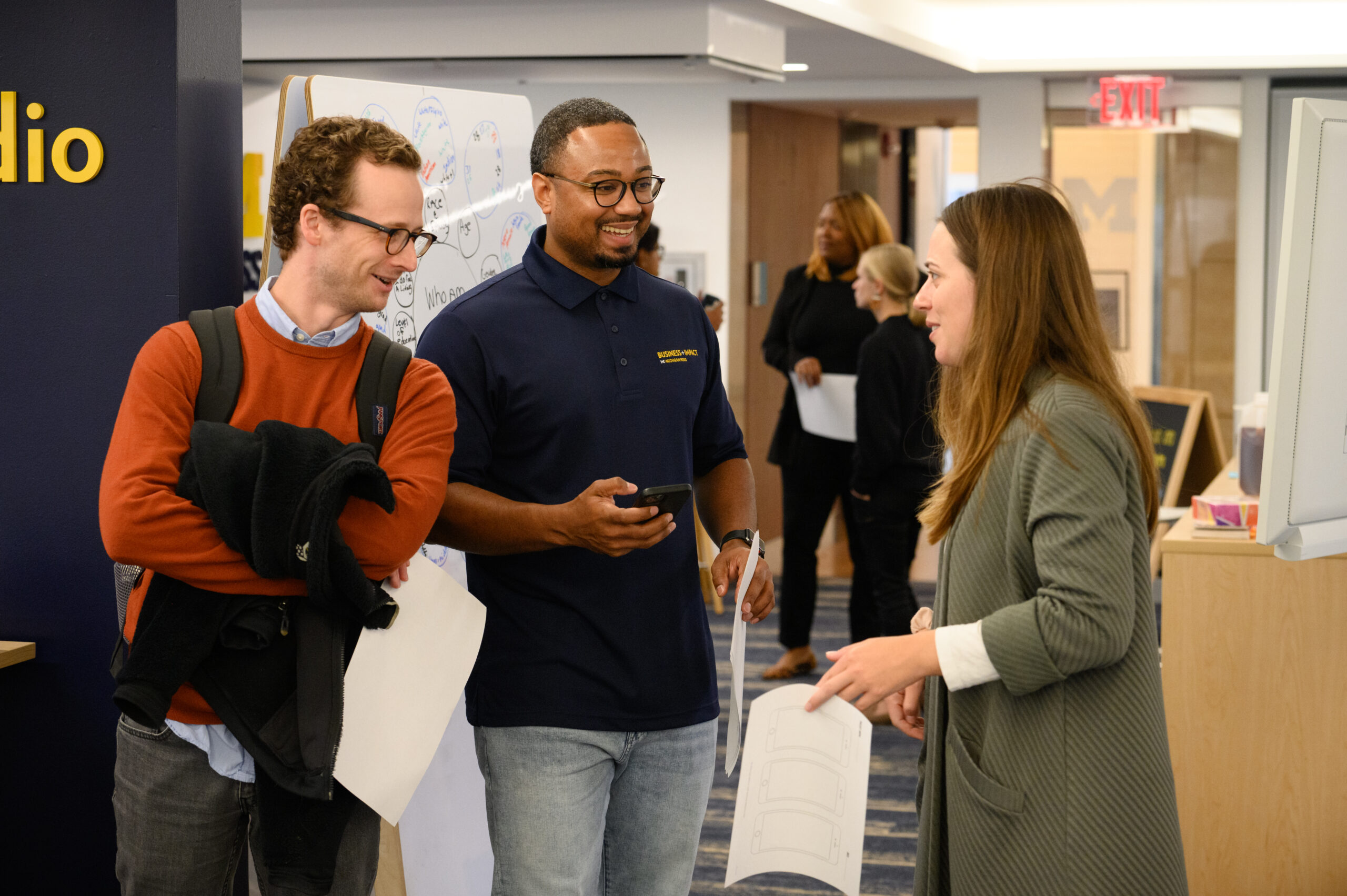 Three colleagues engaged in a lively conversation during a workshop, with one holding a smartphone and another with a paper in hand. They stand near a whiteboard covered in diagrams, fostering a collaborative and engaging atmosphere in the workspace