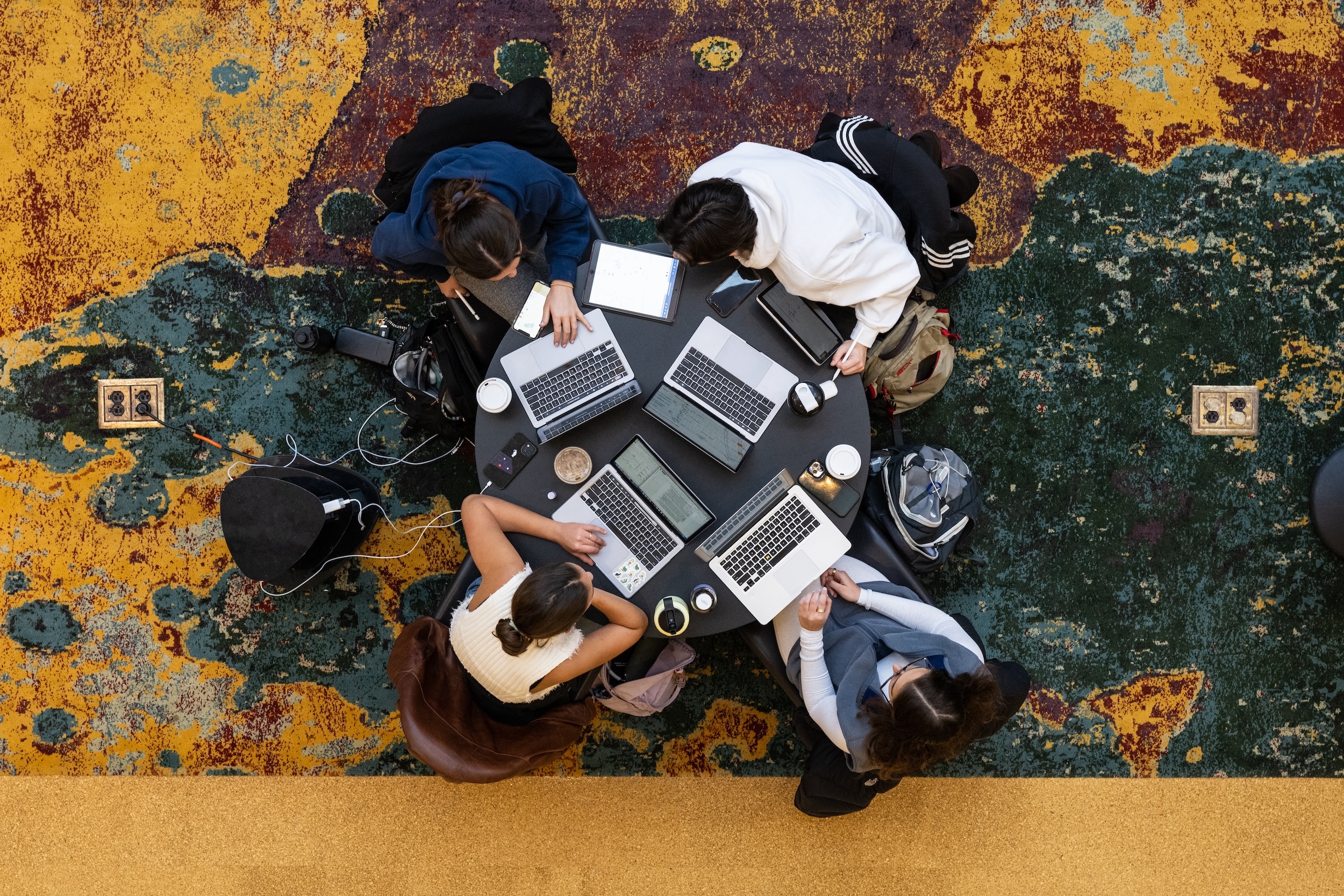 Overhead view of a group of students gathered around a circular table, working on laptops and notebooks. The colorful, textured carpet beneath them adds vibrancy to the scene, as they collaborate and study together in a shared academic space.