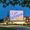 Evening view of a building on the University of Michigan campus featuring a large illuminated display reading 'I am Michigan.' The display glows against the twilight sky, framed by trees and surrounded by softly lit pathways and greenery, creating a welcoming and vibrant campus atmosphere.