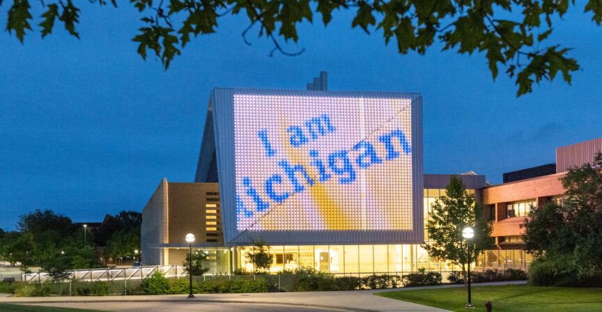 Evening view of a building on the University of Michigan campus featuring a large illuminated display reading 'I am Michigan.' The display glows against the twilight sky, framed by trees and surrounded by softly lit pathways and greenery, creating a welcoming and vibrant campus atmosphere.