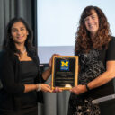 Two individuals stand together during an award presentation. The individual on the left, wearing a black dress and pearl necklace, smiles while holding one side of a wooden plaque. The individual on the right, also smiling, wears a black dress with floral patterns and holds the other side of the plaque. The plaque features the University of Michigan logo and text recognizing an award recipient. A projector screen and curtain serve as the backdrop