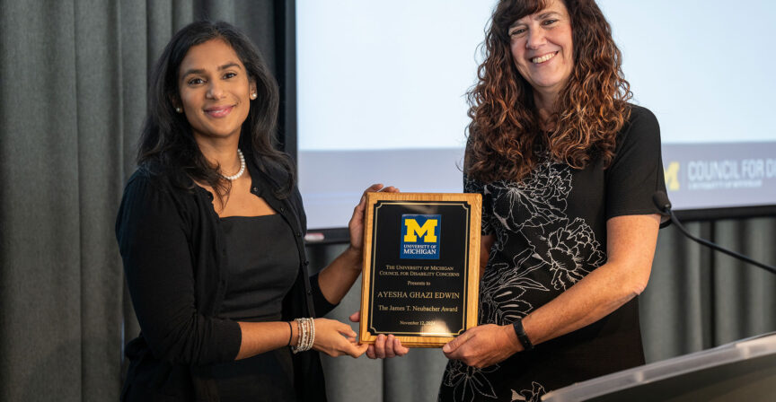 Two individuals stand together during an award presentation. The individual on the left, wearing a black dress and pearl necklace, smiles while holding one side of a wooden plaque. The individual on the right, also smiling, wears a black dress with floral patterns and holds the other side of the plaque. The plaque features the University of Michigan logo and text recognizing an award recipient. A projector screen and curtain serve as the backdrop