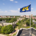 A blue and yellow University of Michigan flag flies prominently above a campus rooftop, with historic buildings, lush green trees, and city structures visible in the background under a bright, clear sky.