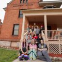 A group of people pose for a photo in front of a historic brick building with the words "ESKOONWID ENDAAD" displayed at the top. The individuals are gathered on and around the porch stairs.